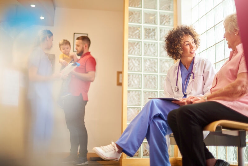 A female nurse sits in doctor’s waiting room with senior female patient taking notes and updating her medical records, another patient checks his details are correct with another nurse in the background